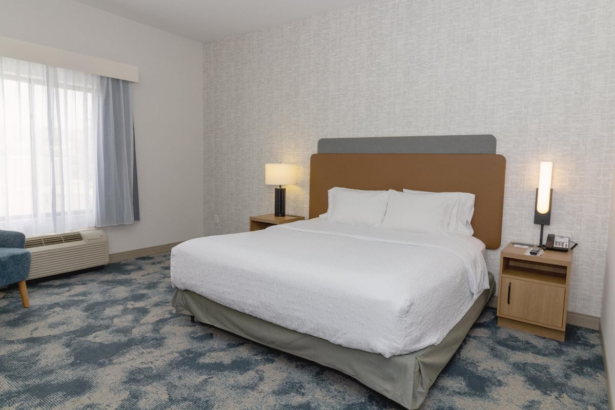 A hotel room with a large bed, two bedside tables with lamps, a window with curtains, and a wall-mounted air conditioner.