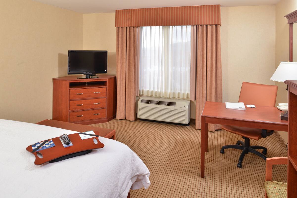 A hotel room with a bed, a desk, a chair, a television, a dresser, and an air conditioning unit.