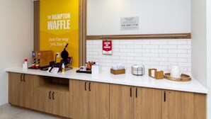 Uma área de recepção de hotel com balcão, armários de madeira, um letreiro amarelo para The Hampton Waffle e um aviso de cuidado com superfícies quentes.