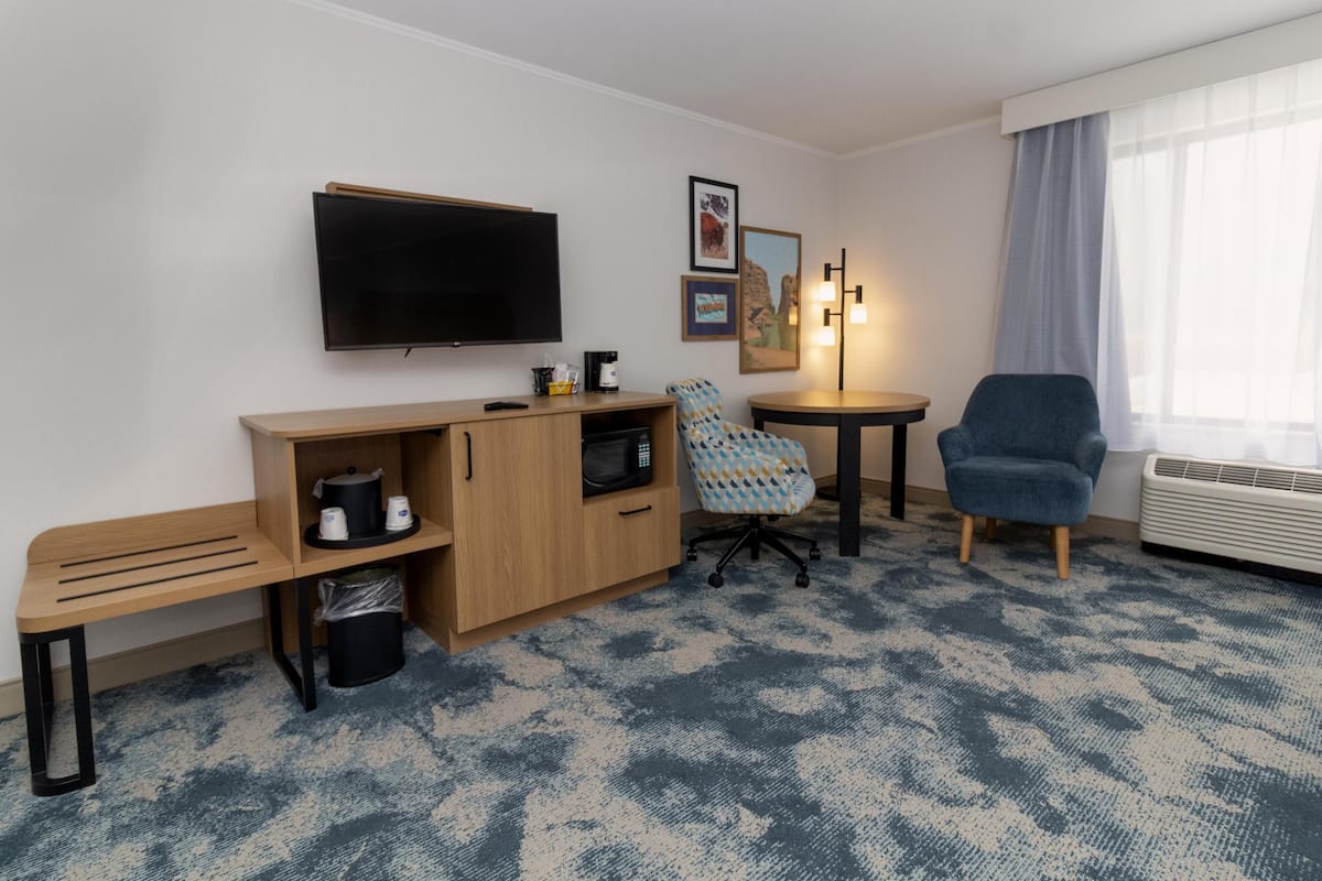 A hotel room with a flat-screen TV, a wooden desk, a blue patterned armchair, a blue armchair, a small table, and a window with curtains.