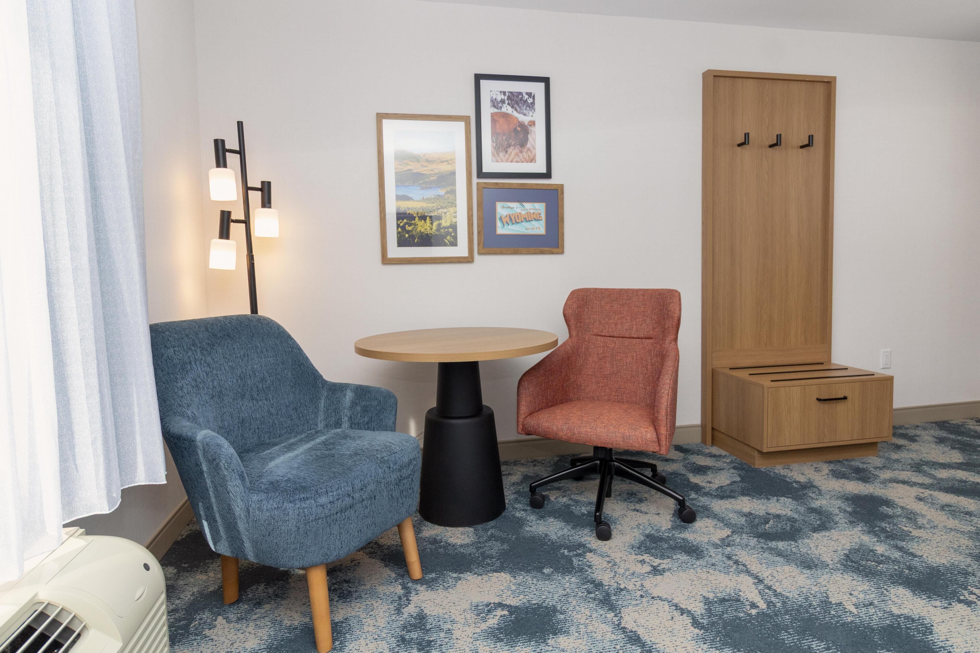 Um quarto com uma poltrona azul, uma poltrona vermelha, uma mesa redonda, um abajur, um guarda-roupa e três quadros na parede.