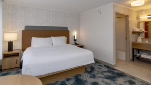 Quarto de hotel com cama, mesa de cabeceira, luminária e banheiro com espelho e porta-toalhas.