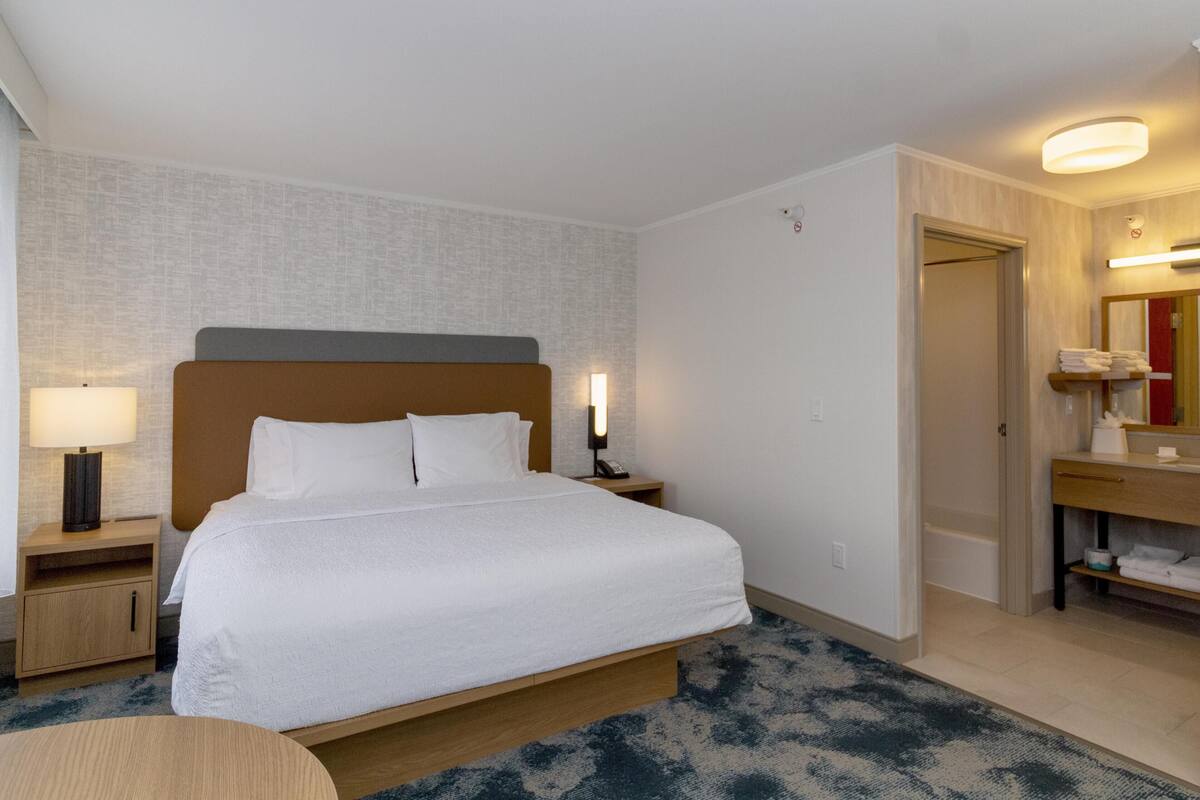 A hotel room with a bed, bedside table, lamp, and a bathroom with a mirror and towel rack.