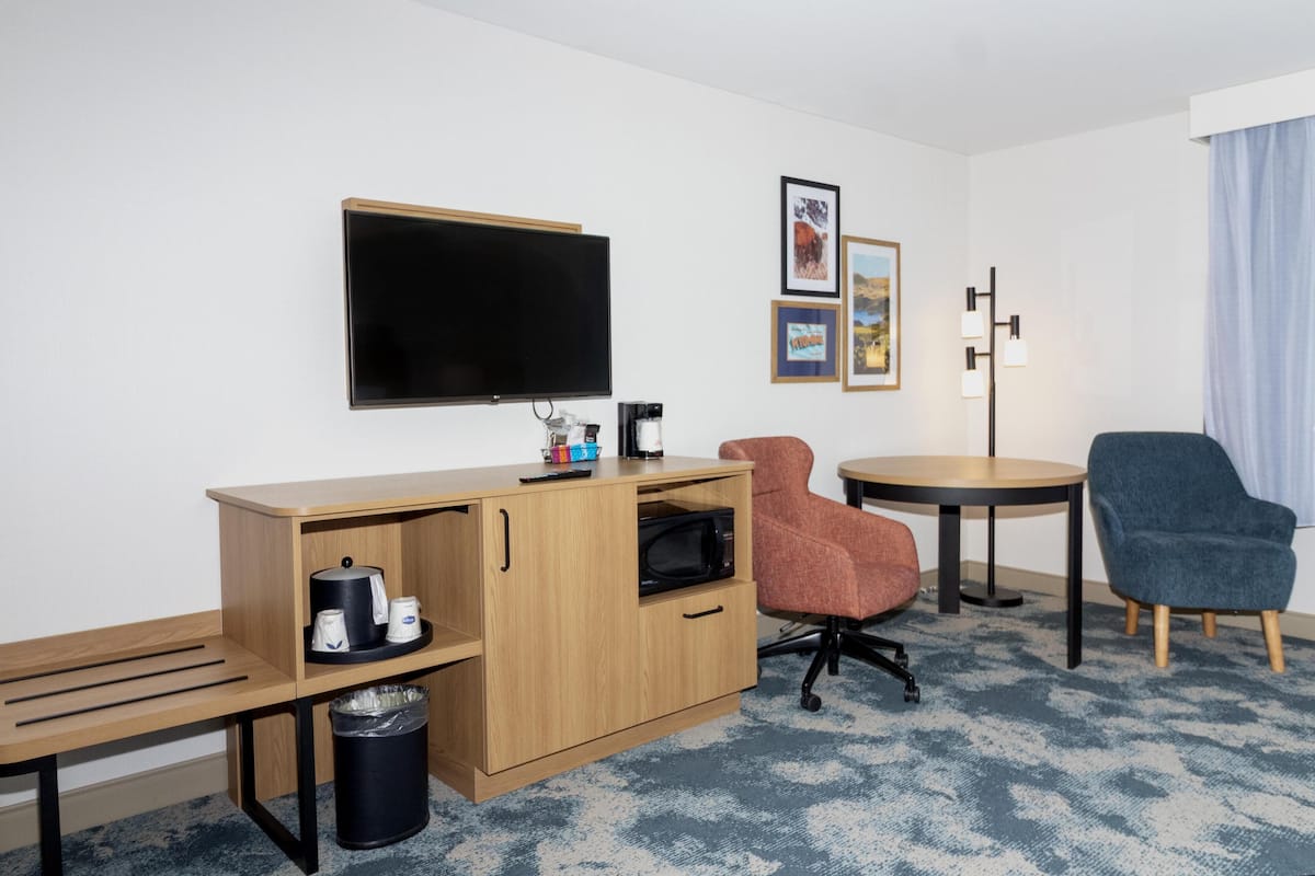 A hotel room with a wooden desk, a television, a coffee maker, a trash can, two chairs, a small table, and framed pictures on the wall.