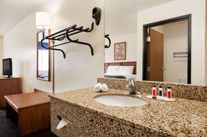 Baño de hotel con lavamanos de encimera de granito, toallero, espejo y televisor sobre una mesita de madera.
