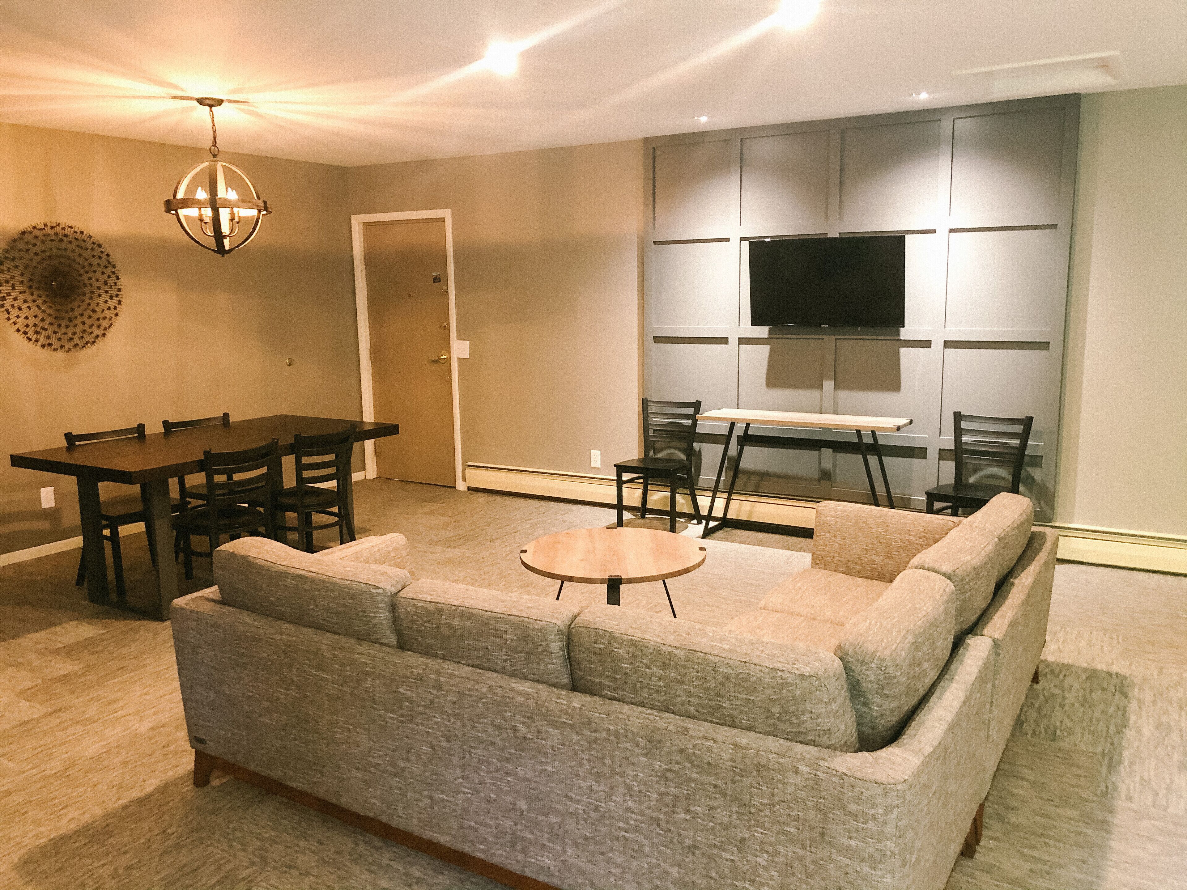A living room with a sectional sofa, a dining table with chairs, a small round table, a ceiling fan, and a flat-screen TV mounted on the wall.