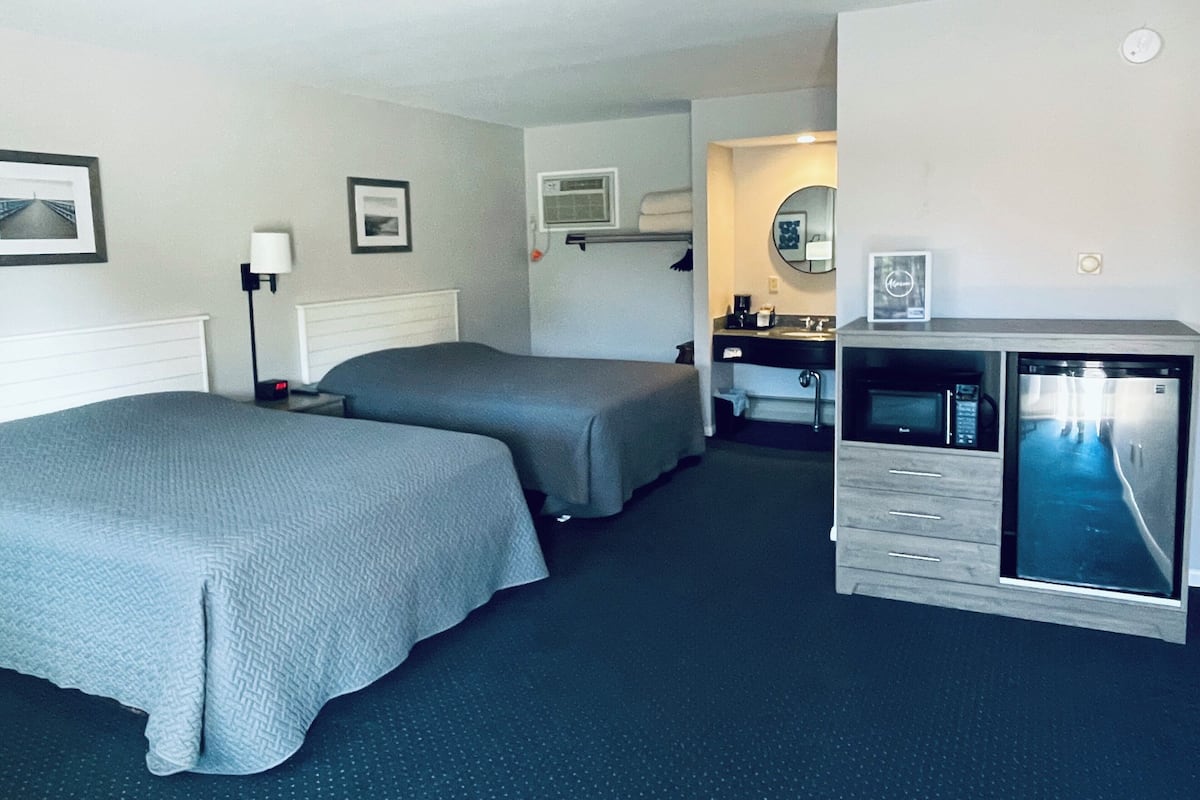 A hotel room with two single beds, a desk with a computer, a small kitchenette, and a bathroom with a mirror and sink.