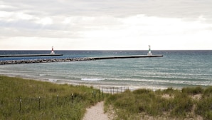 Escena costera con dos faros en un muelle que se adentra en el mar, una playa arenosa y un sendero de hierba que lleva al agua.