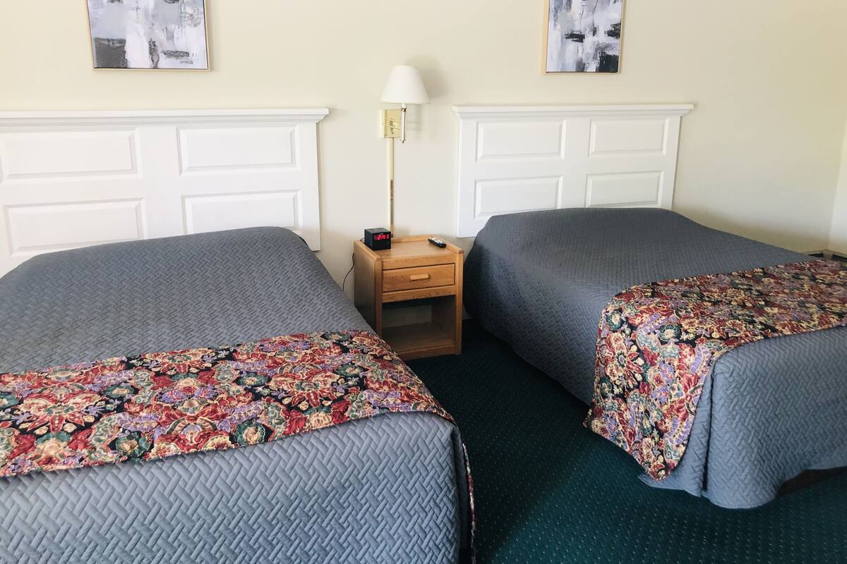 A hotel room with two beds, each with a grey quilt and a floral bedspread. There is a wooden nightstand with a lamp and a clock between the beds. The walls are plain with two abstract paintings above the beds.