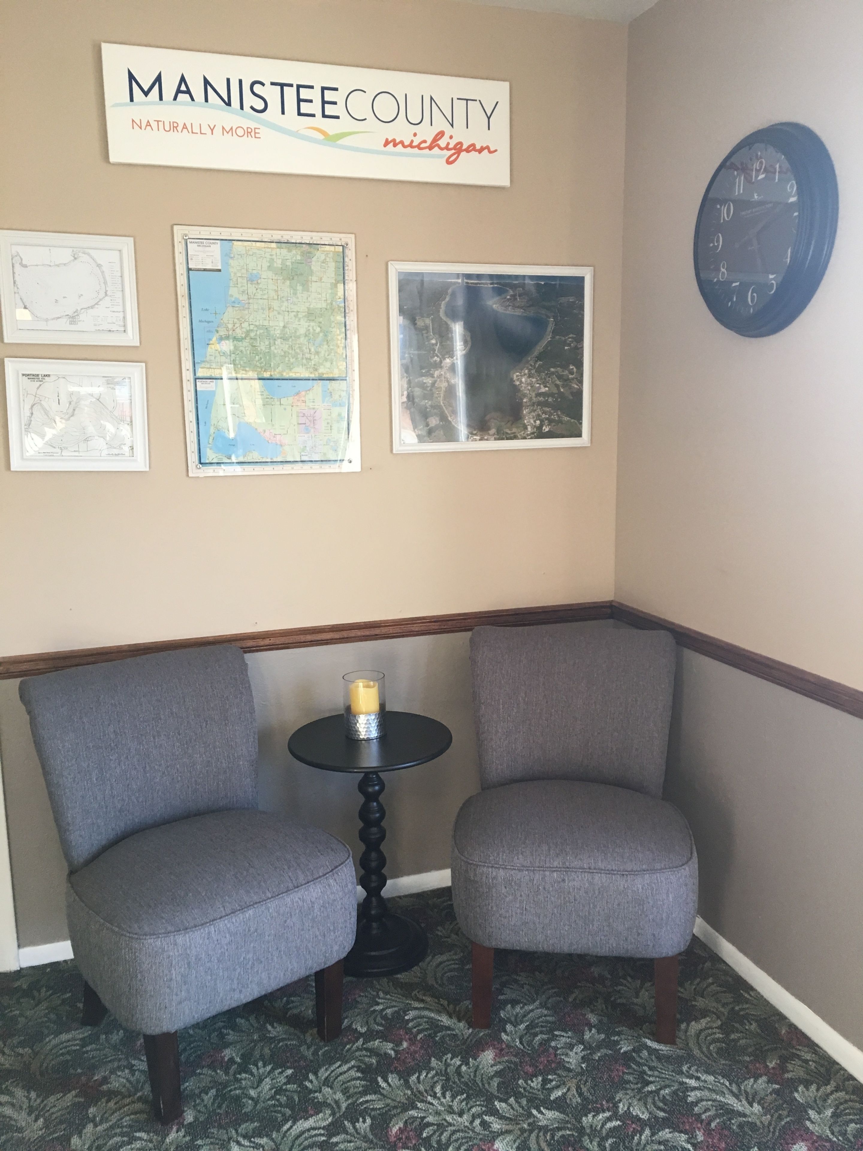 A corner room with two grey armchairs, a small table with a candle, framed maps, and a clock on the wall.
