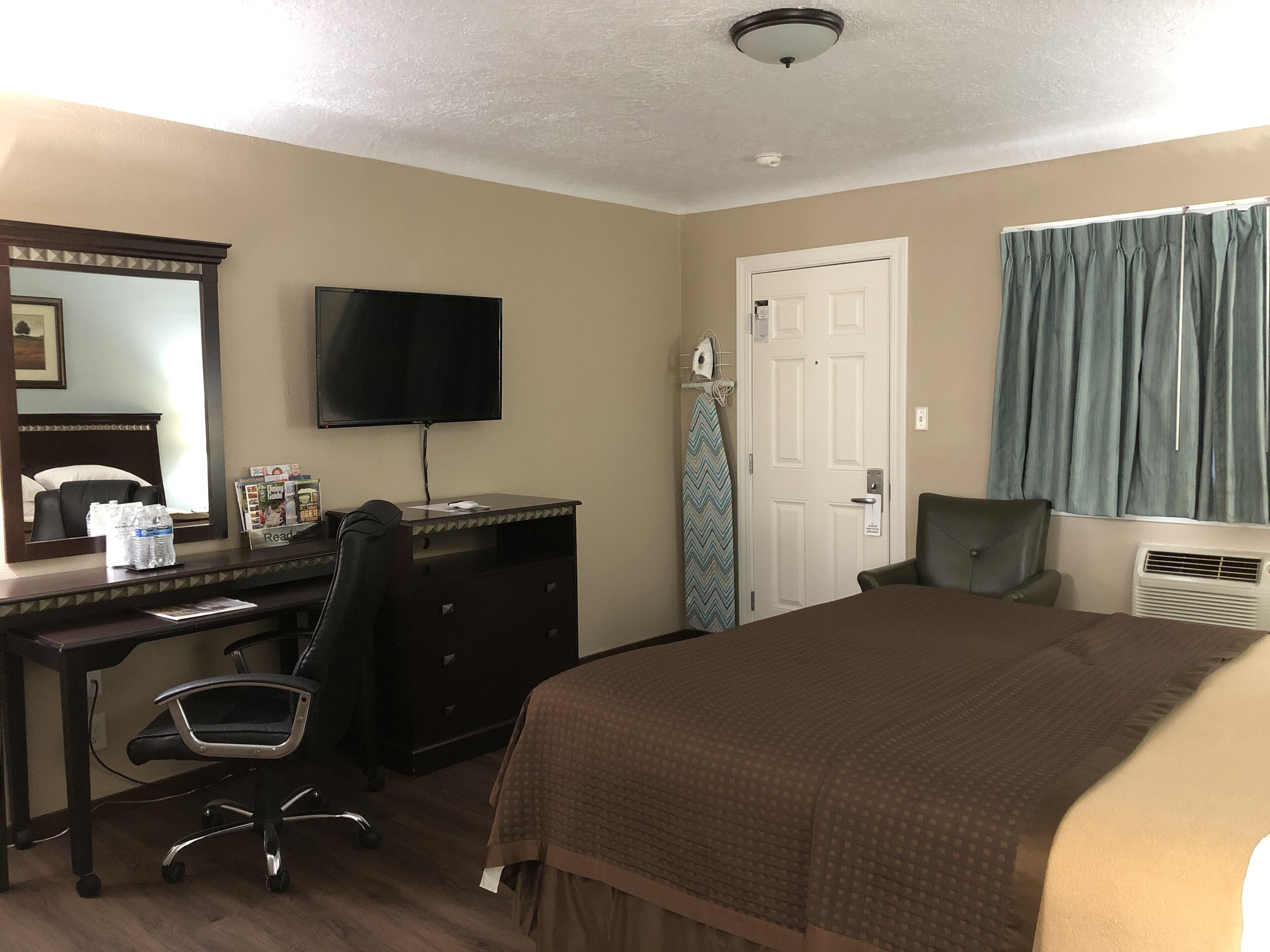 Une chambre d’hôtel comprenant un lit, un bureau avec une chaise, une télévision, une porte et une fenêtre avec des rideaux.