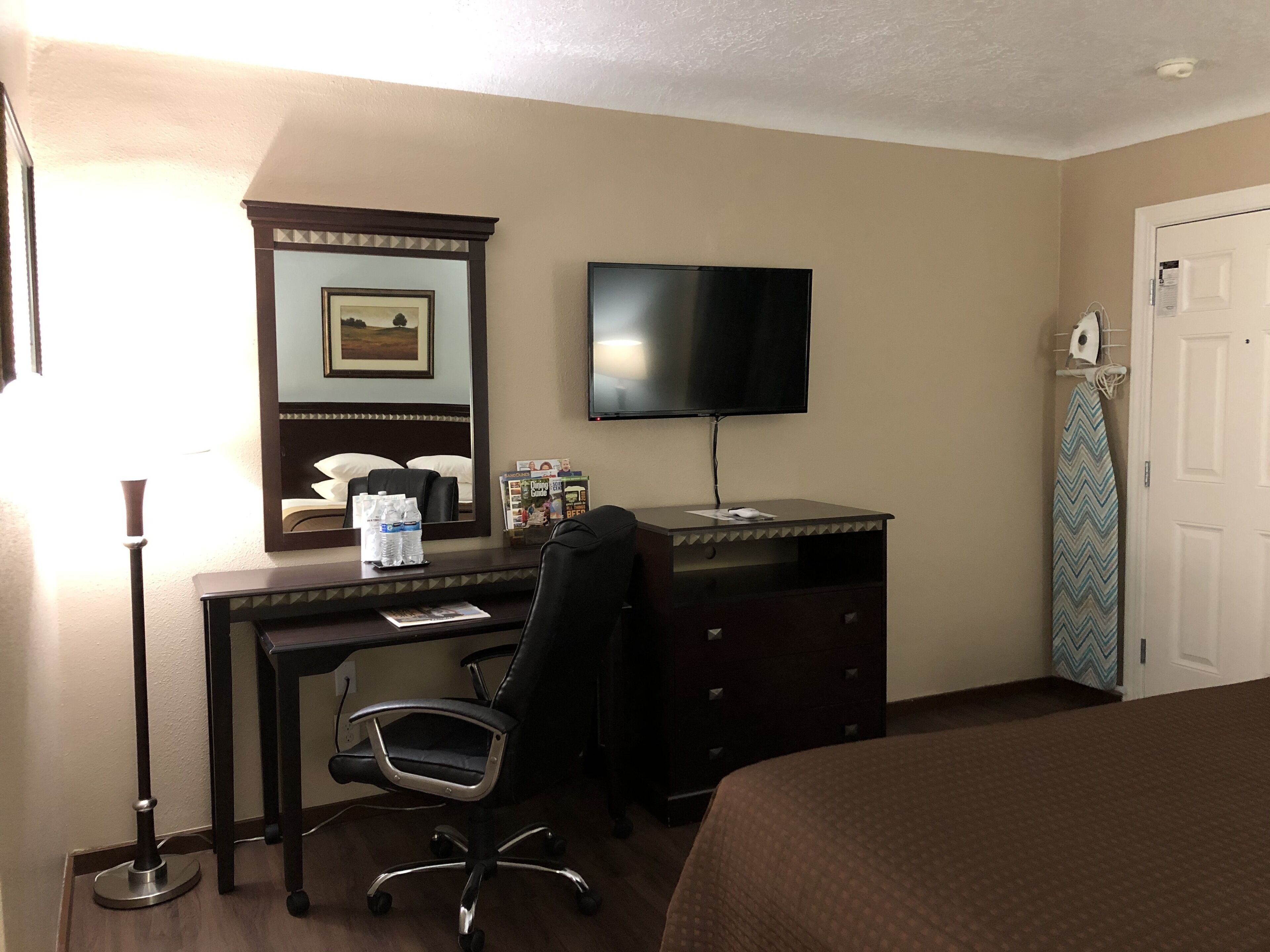 A hotel room with a bed, a desk with a chair, a flat-screen TV, a dresser, a mirror, and a door.