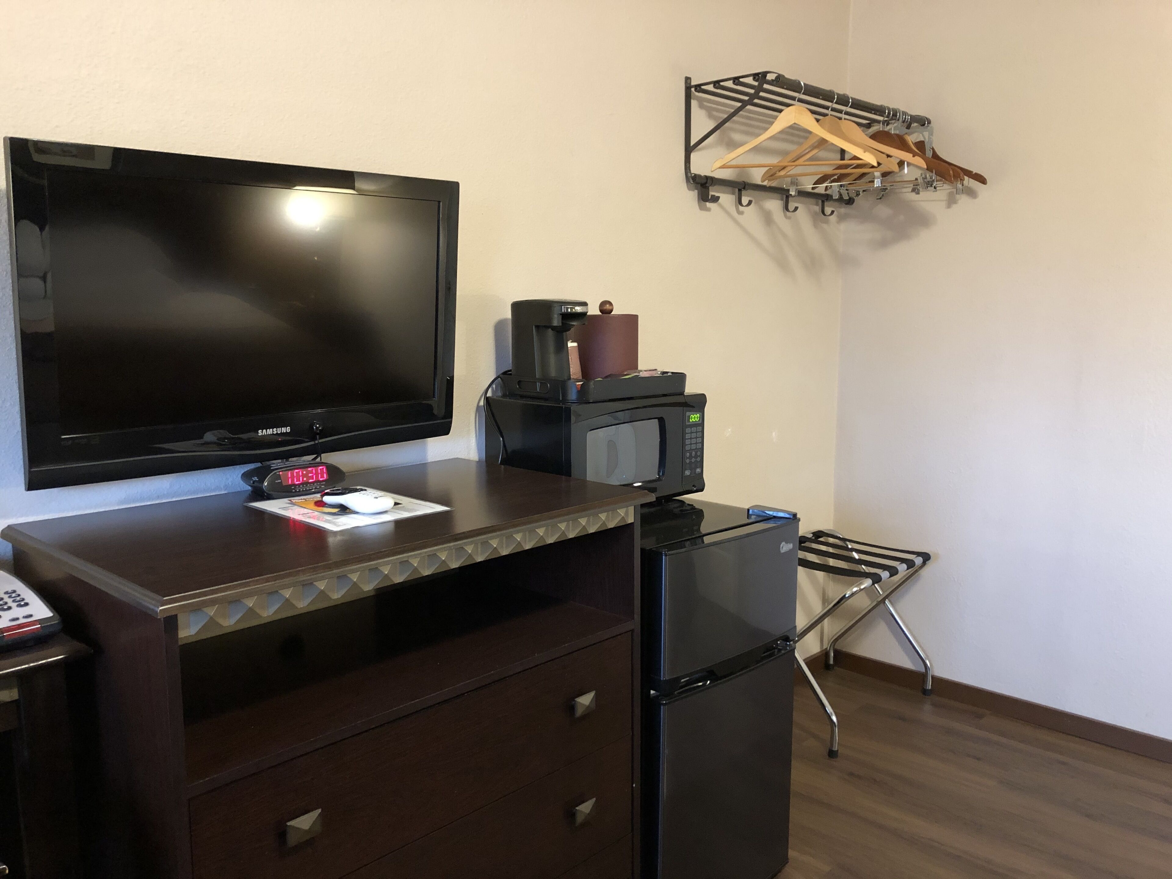 A hotel room with a flat-screen TV, a coffee maker, a microwave, a refrigerator, a coat rack, and a small table with a clock and a remote control.