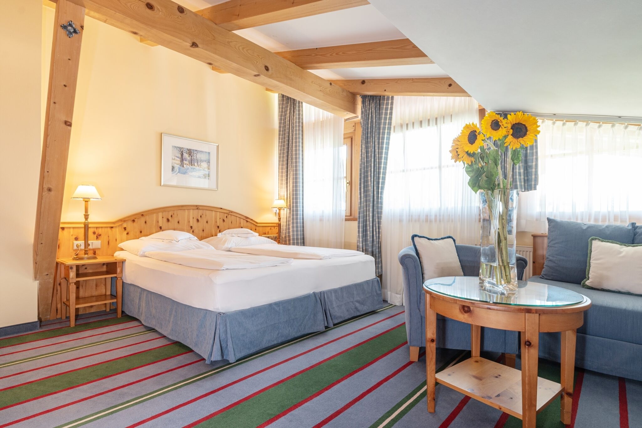 Habitación de hotel con una cama, una mesita con un jarrón de girasoles, un sofá y una ventana con cortinas.