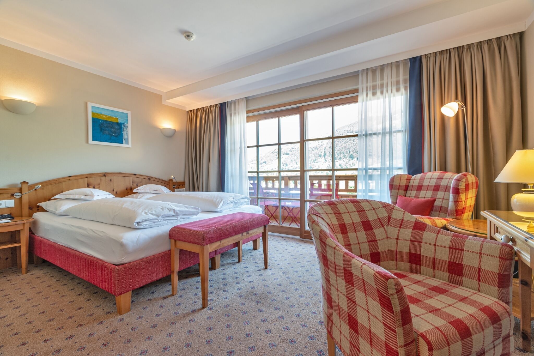 A hotel room with a double bed, a bench, a plaid armchair, a table with a lamp, and a large window with curtains.