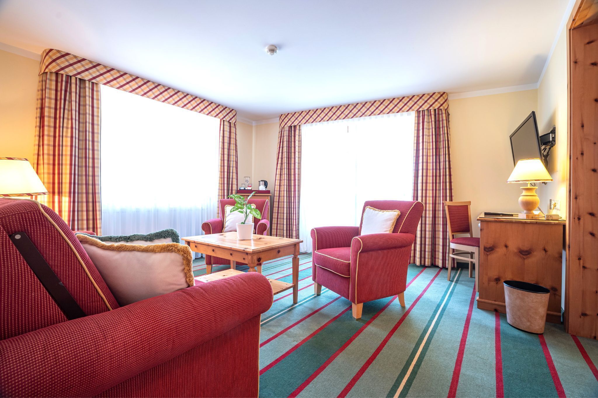 Habitación de hotel con un sofá rojo, un sillón rojo, una mesita de madera, una pequeña mesa con una planta y un armario de madera.