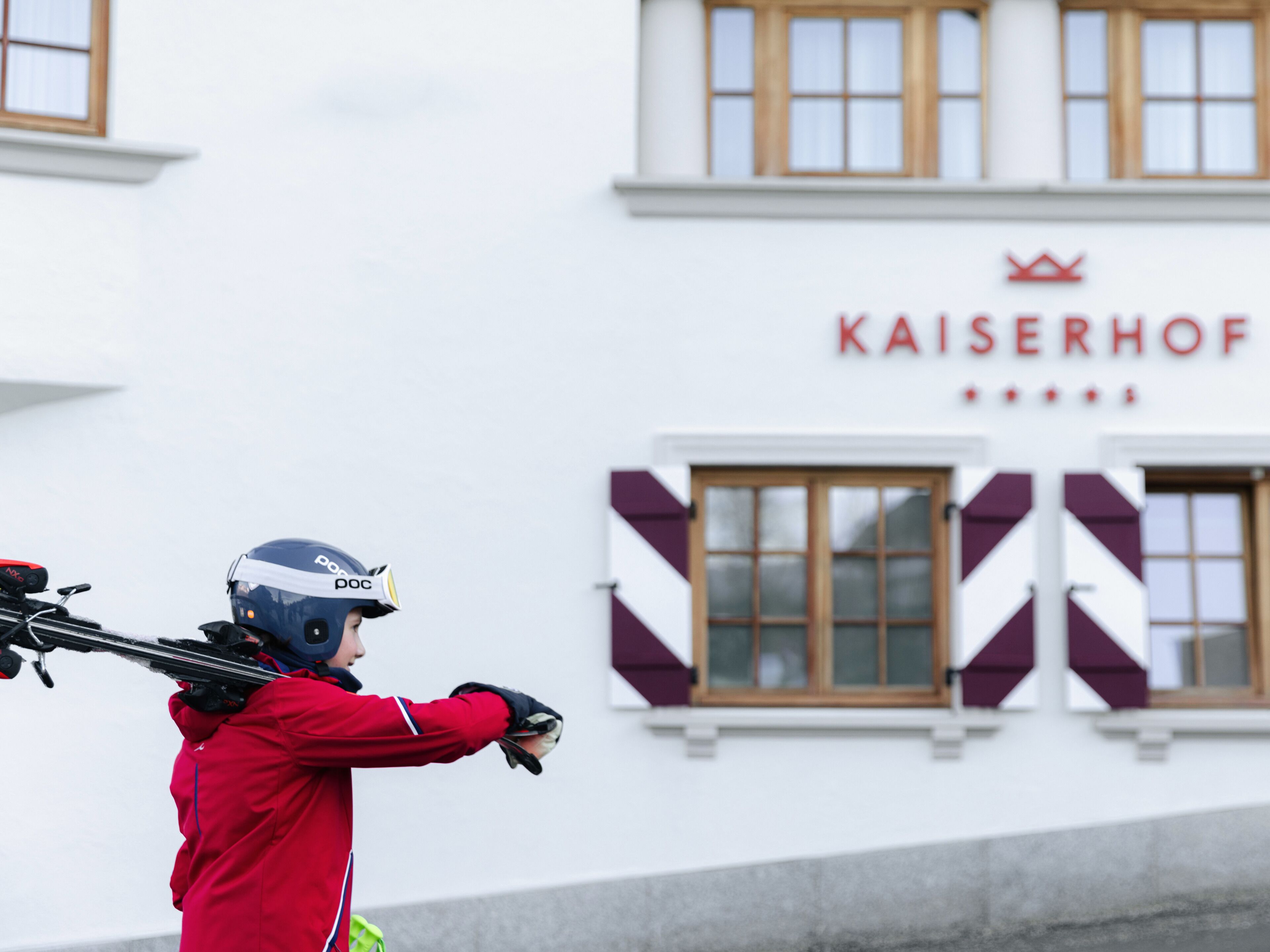 Een persoon in een rode jas met helm, ski's vasthoudend voor een gebouw met het bord 'KAISERHOF'.