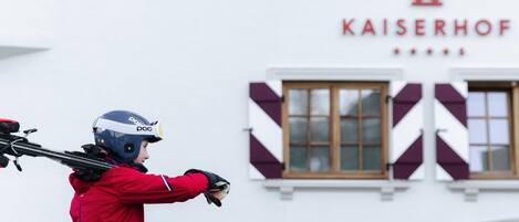 A person in a red jacket with a helmet, holding skis in front of a building with the sign 'KAISERHOF'.