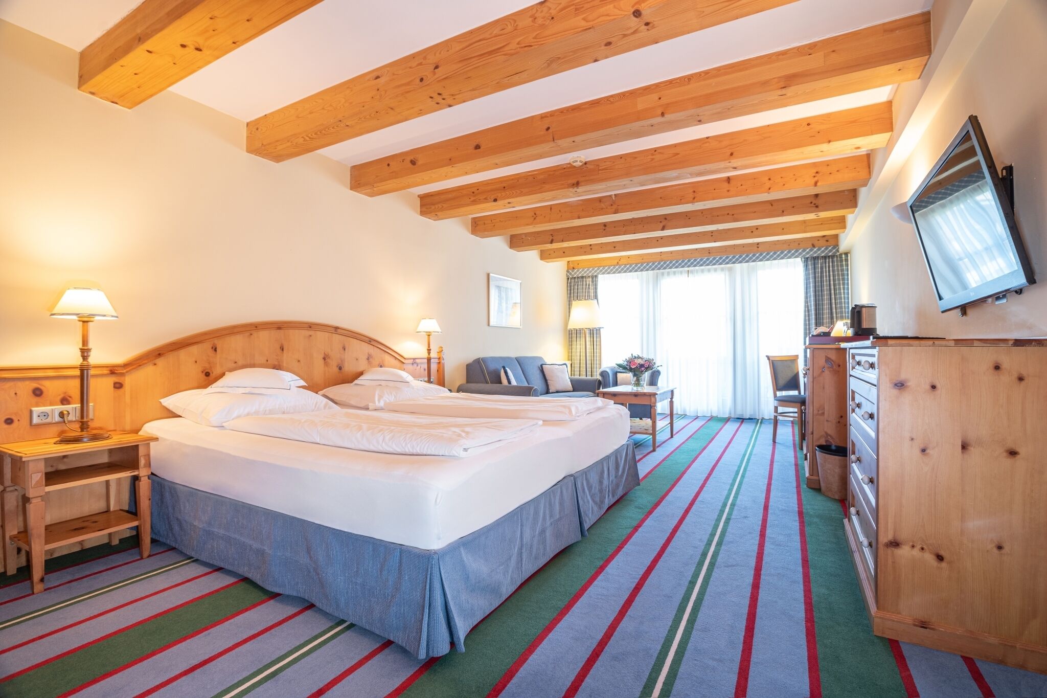 A hotel room with a wooden ceiling, a bed with white linens, a small table with a lamp, a chair, and a TV mounted on the wall.