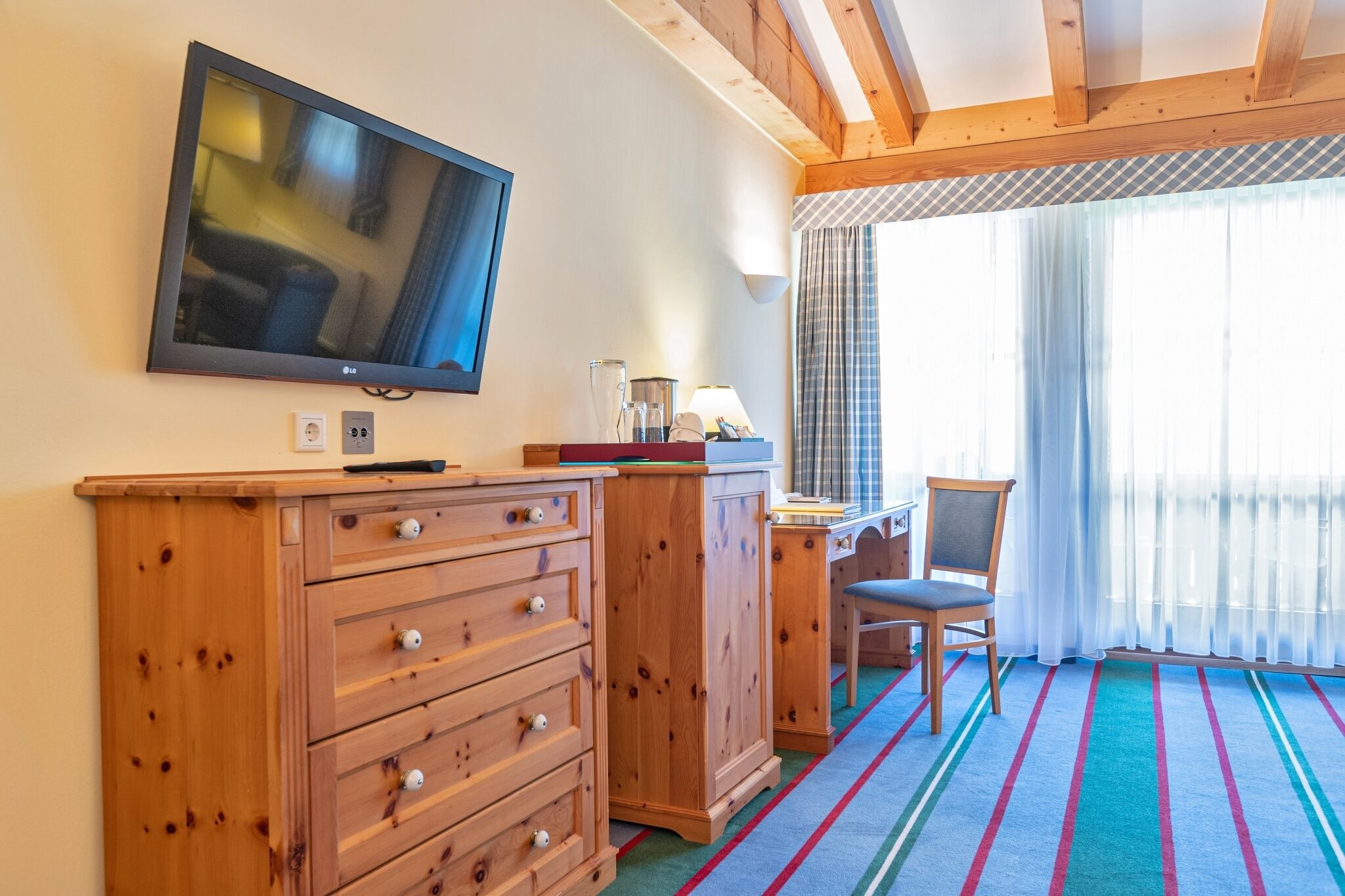Quarto com televisão, cómoda de madeira, secretária, cadeira e janela com cortinas.