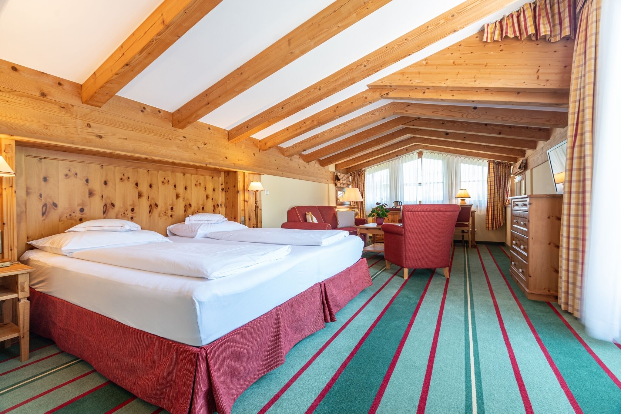 Una habitación con techo de madera, una cama con ropa blanca, una silla roja y un escritorio con lámpara.