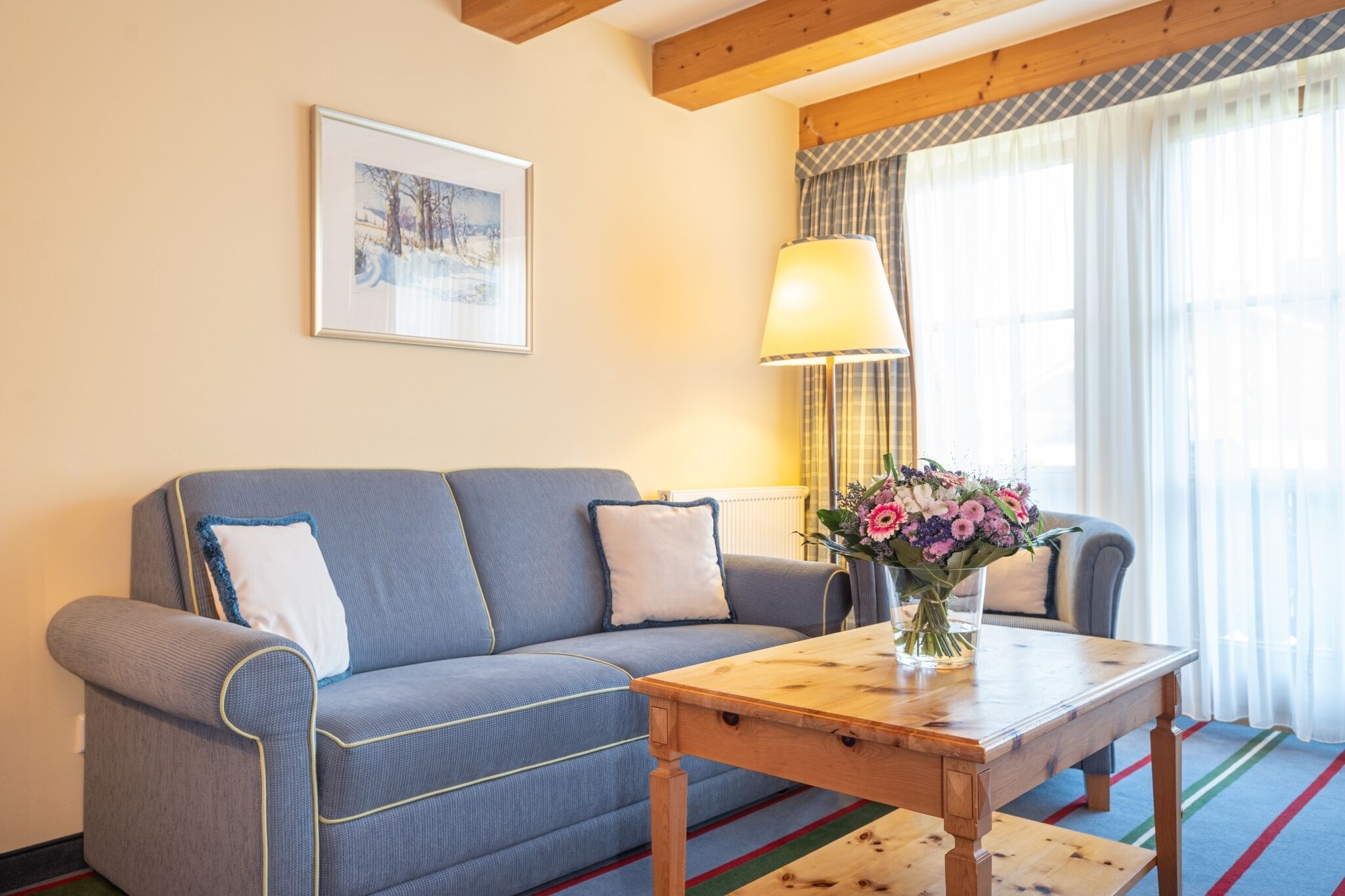 Um sofá azul com almofadas brancas, uma mesa de centro de madeira com um vaso de flores, um quadro na parede e uma janela com cortinas.
