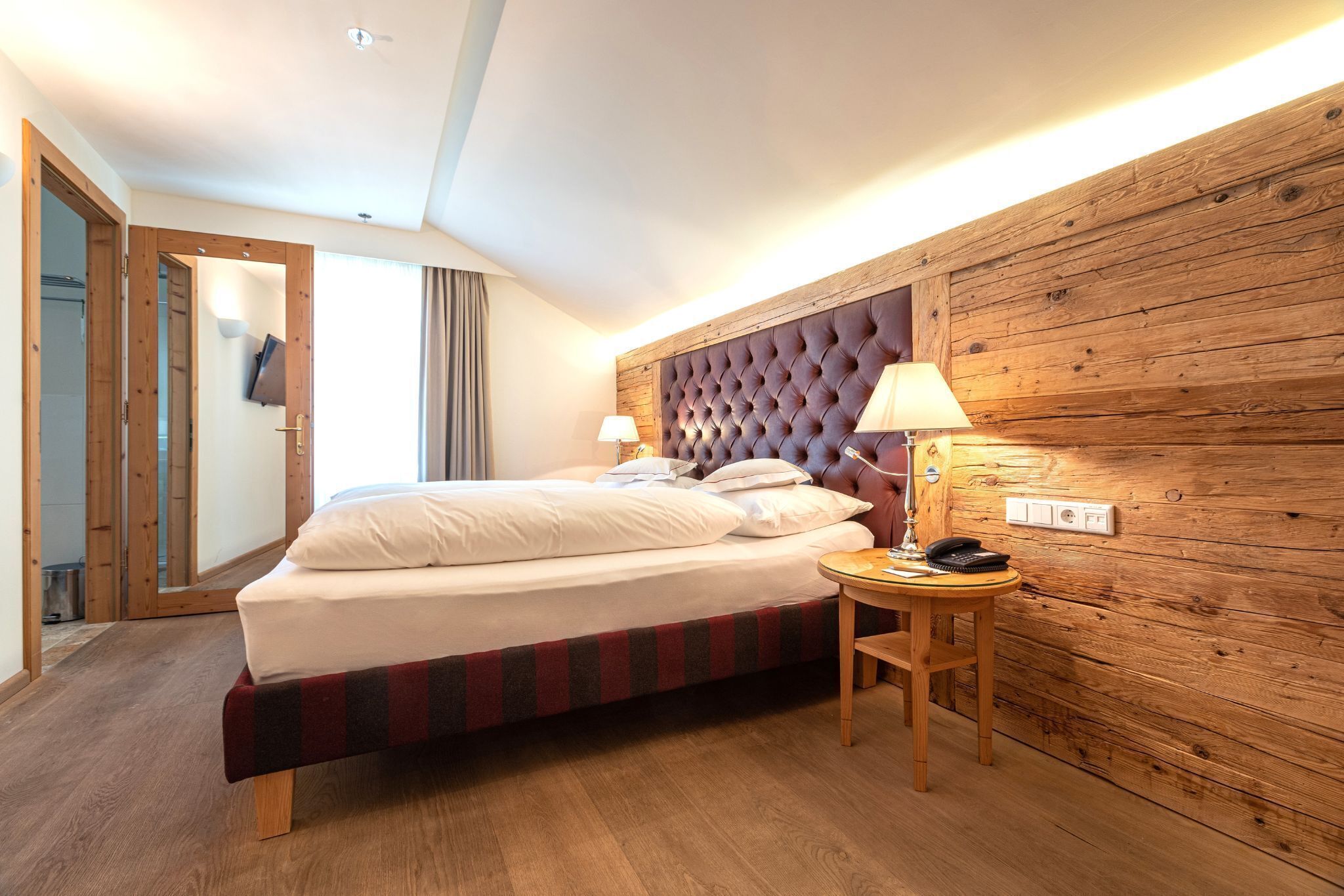 A hotel room with a bed, wooden headboard, bedside table, lamp, and a view of the outside.