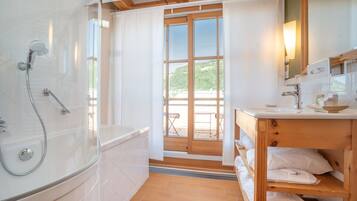 Baño con bañera, ducha, tocador de madera y vistas a una terraza y vegetación.
