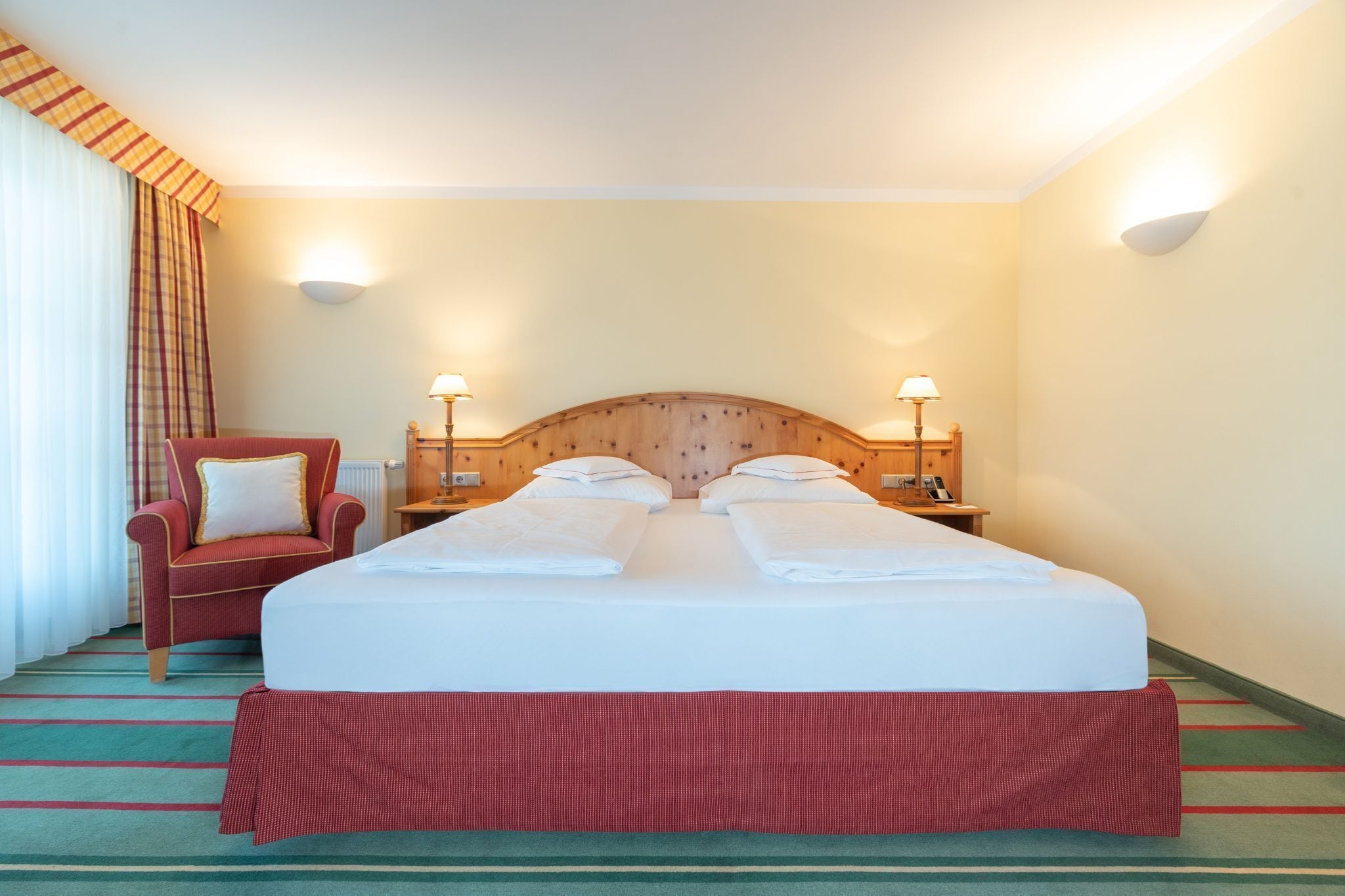 Ett hotellrum med en dubbelsäng, två sängbord med lampor, en röd fåtölj och en randig matta.