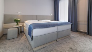 Ett modernt hotellrum med en stor säng, ett litet sidobord med en vas och en grå soffa.