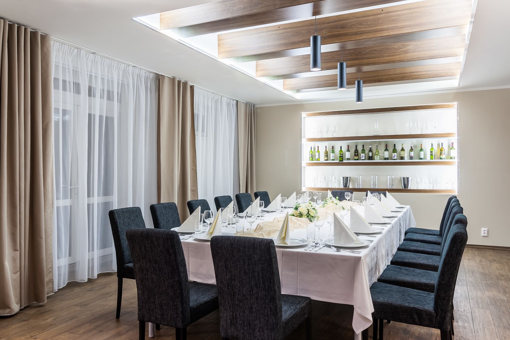 A dining area with a long table set for a meal, surrounded by chairs, with a wine rack and glassware in the background.