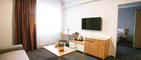 Habitación de hotel con sofá, una mesita con una botella y fruta, un escritorio con una silla, televisor en la pared y un armario.