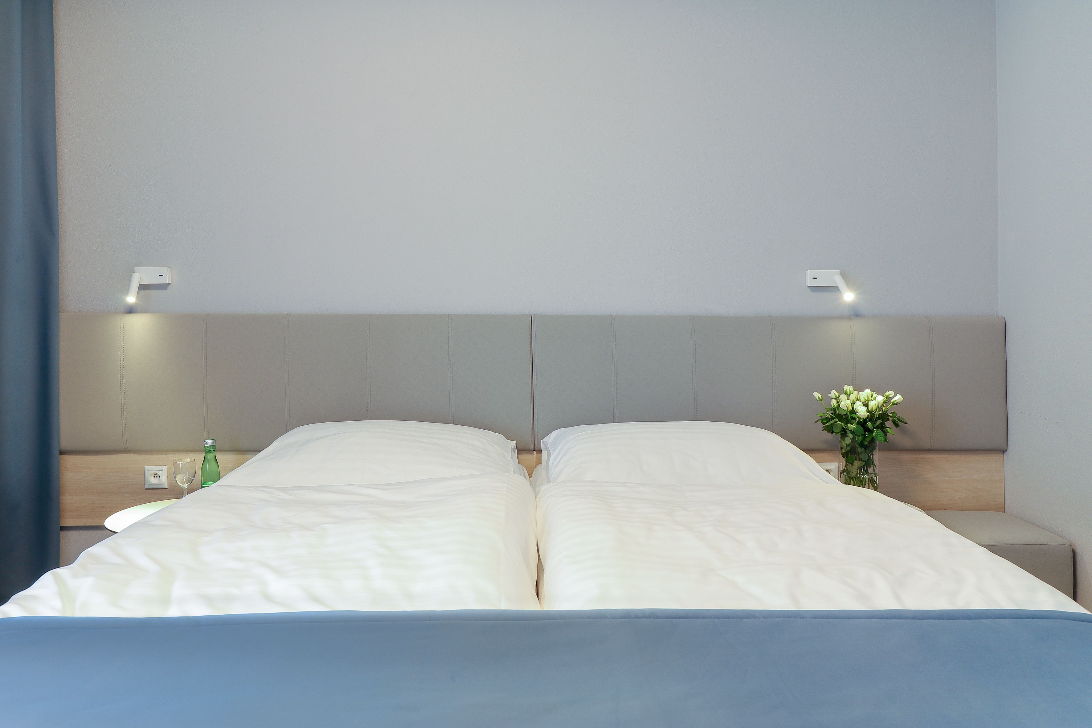 Una cama doble con ropa blanca, una mesita de noche con un vaso de agua y una botella, y un jarrón con flores.