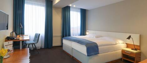 A hotel room with a bed, bedside table, lamp, desk with chair, and a TV.