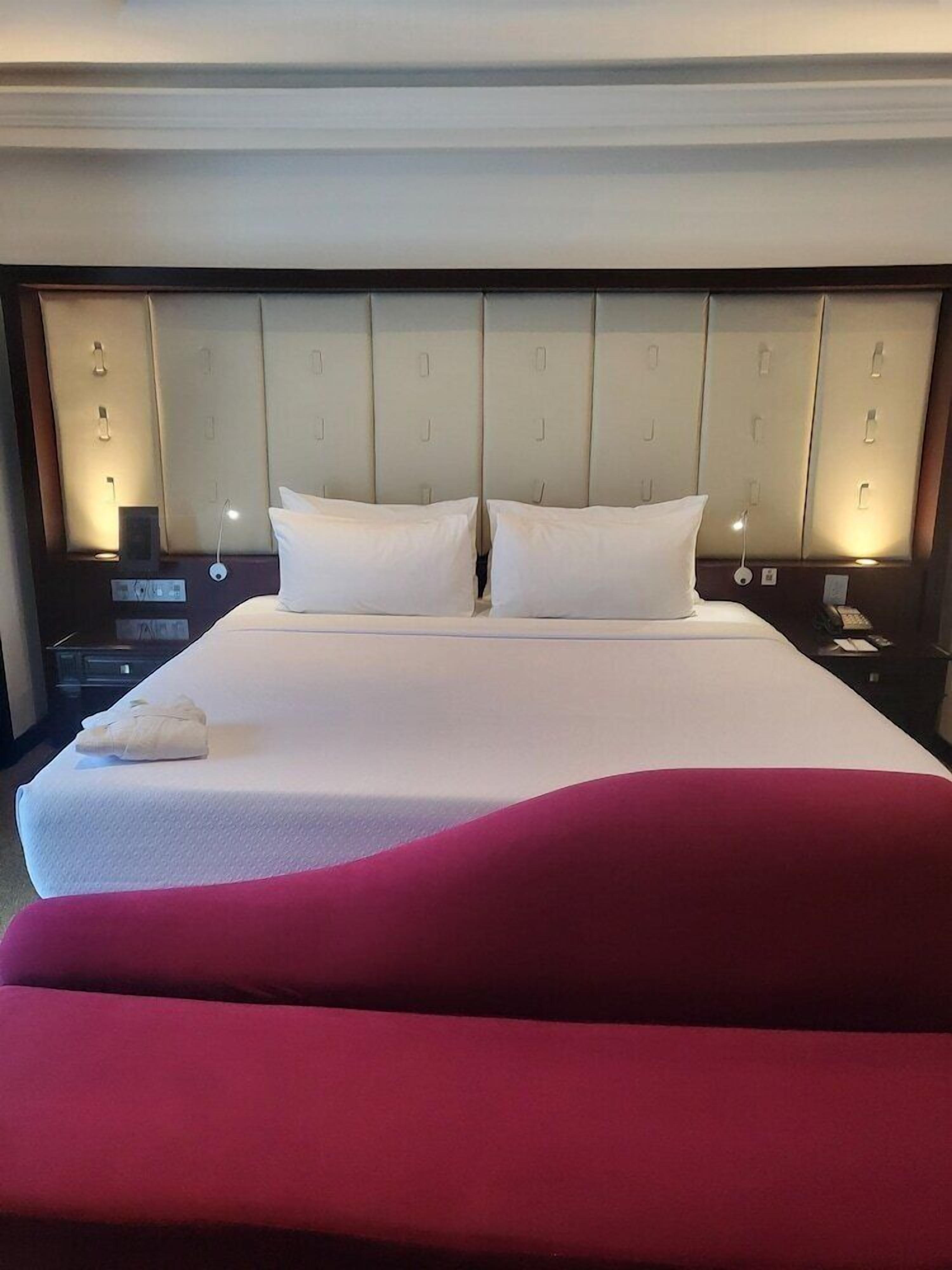 Una habitación de hotel con una cama grande, un cabecero con iluminación integrada, una mesita de noche con lámpara y un detalle rojo.