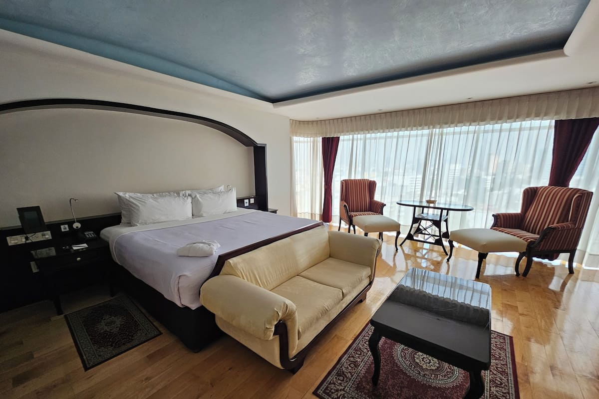 Habitación de hotel con una cama, dos sillones, un sofá, una mesita, una mesita de noche y un ventanal con cortinas.