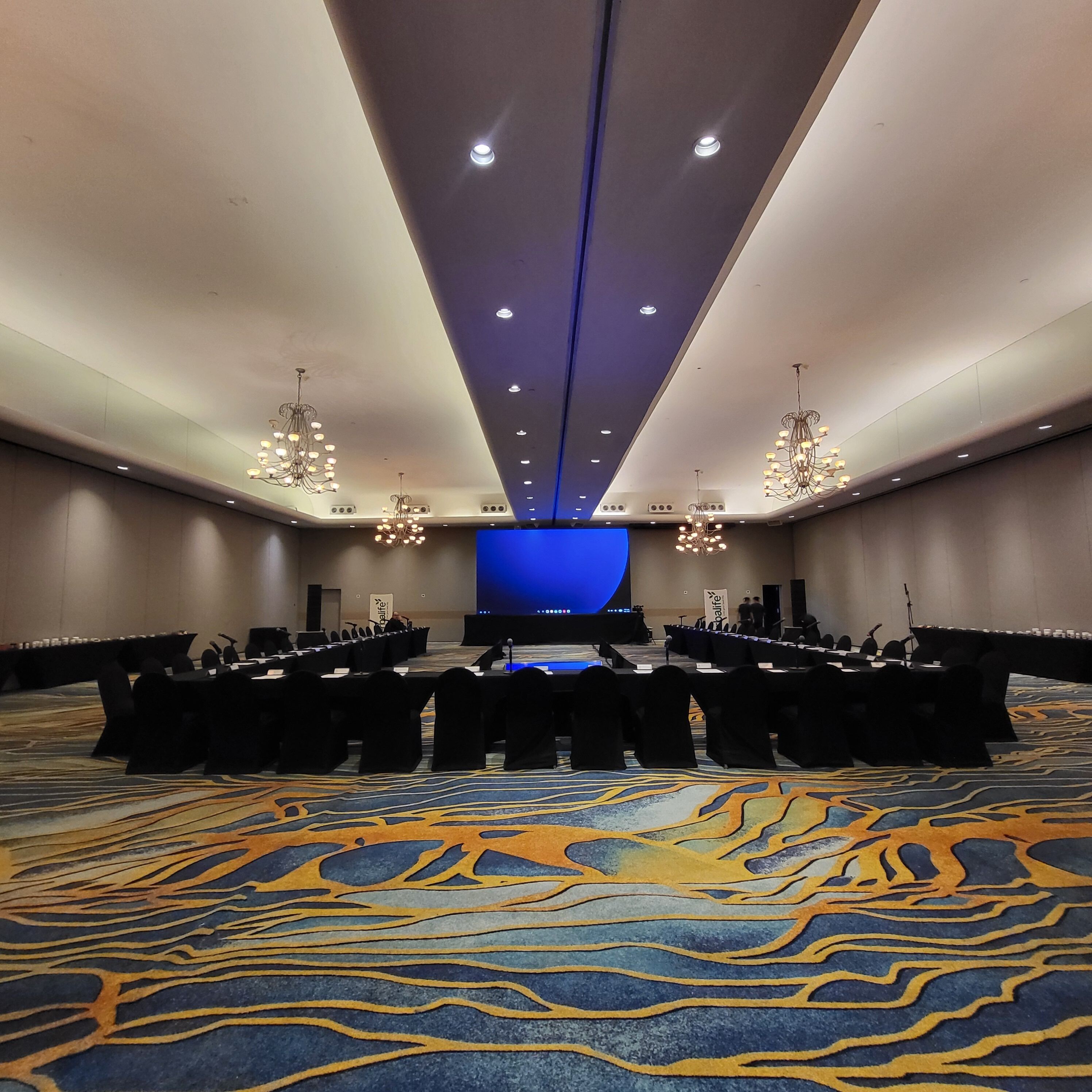 A conference room with a stage, multiple rows of chairs, and chandeliers.