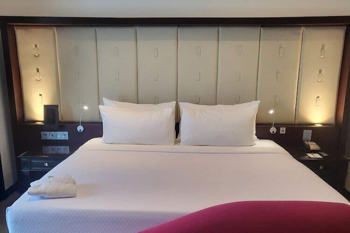 Una habitación de hotel con una cama grande, un cabecero con iluminación integrada, una mesita de noche con lámpara y un detalle rojo.