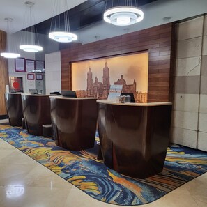 Un área de recepción con diseño moderno, que incluye un mostrador de recepción, varias sillas, una alfombra estampada y un mural con vistas a la ciudad.