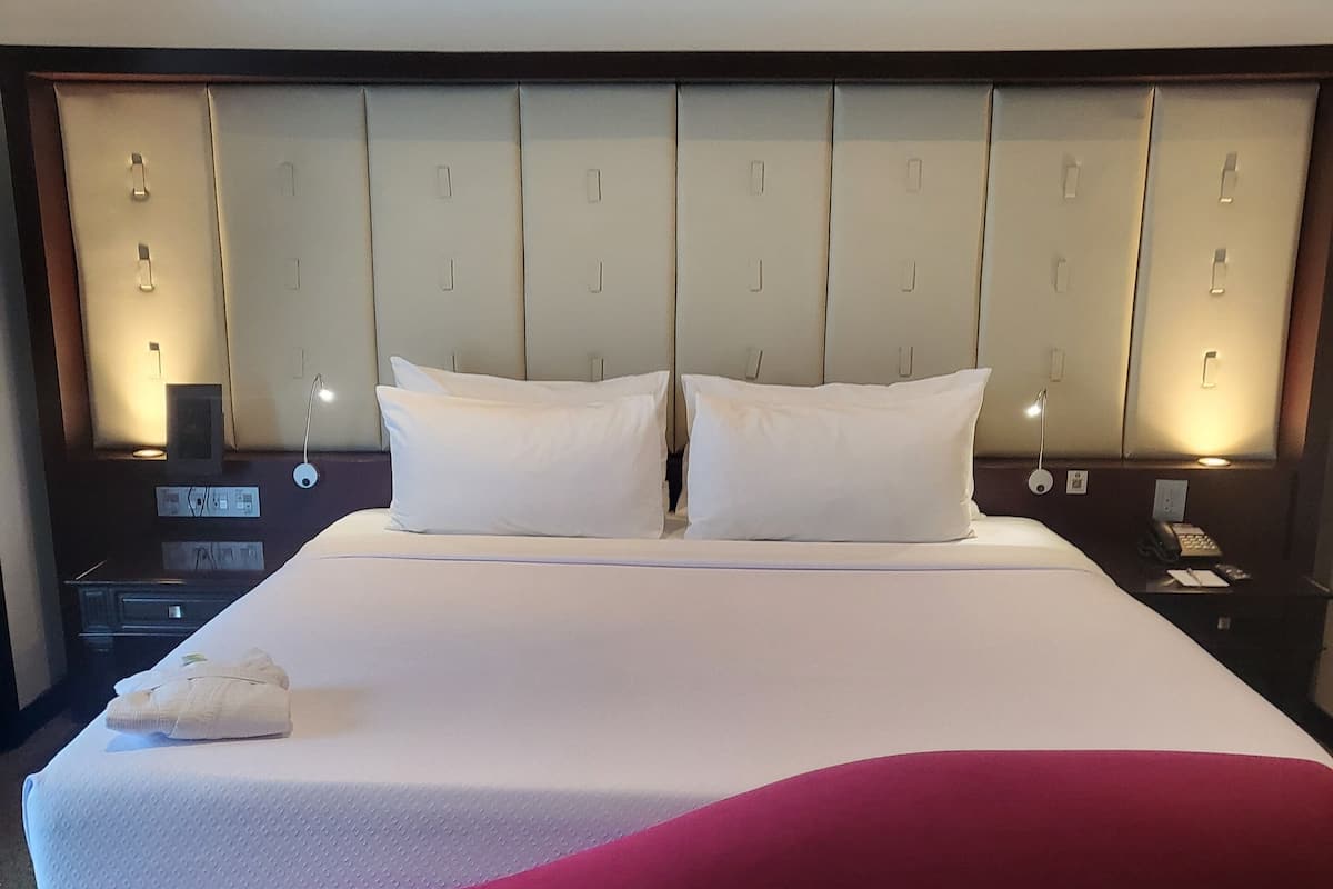 Habitación de hotel con una cama grande, cabecera con iluminación integrada, mesita de noche con teléfono y un cojín decorativo rojo.