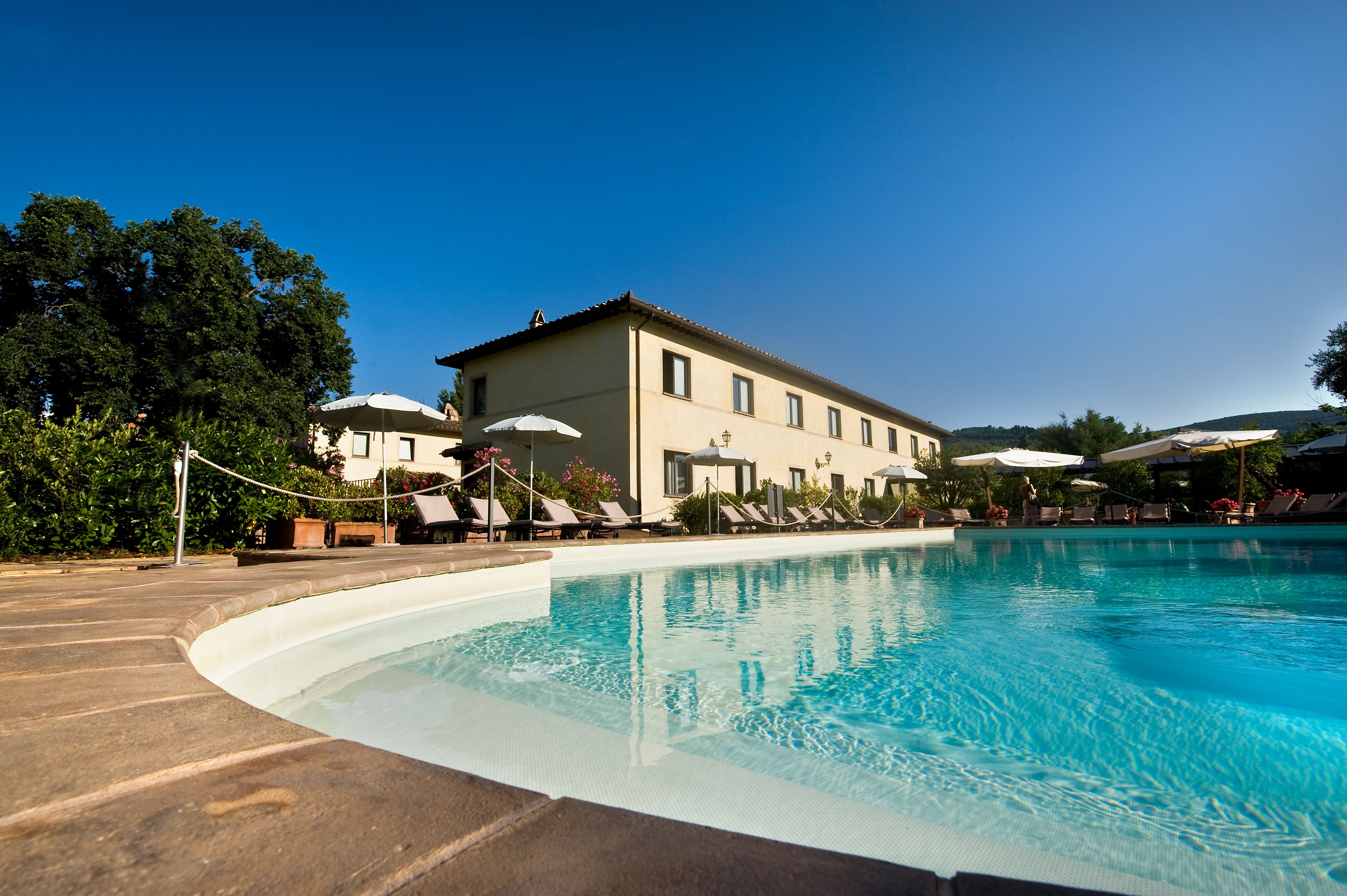 A swimming pool with lounge chairs and umbrellas, a building with a tiled roof, and greenery in the background.