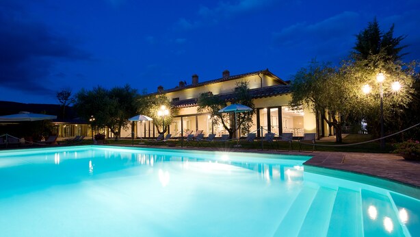 A swimming pool with lounge chairs and umbrellas, surrounded by trees and a building with outdoor lighting. - Relais dell'Olmo (Perugia)