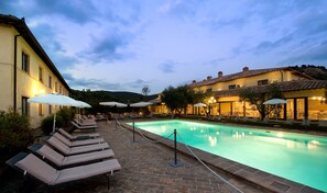 A poolside area with lounge chairs and umbrellas, a brick pool, and buildings with warm lighting. - Relais dell'Olmo (Perugia)