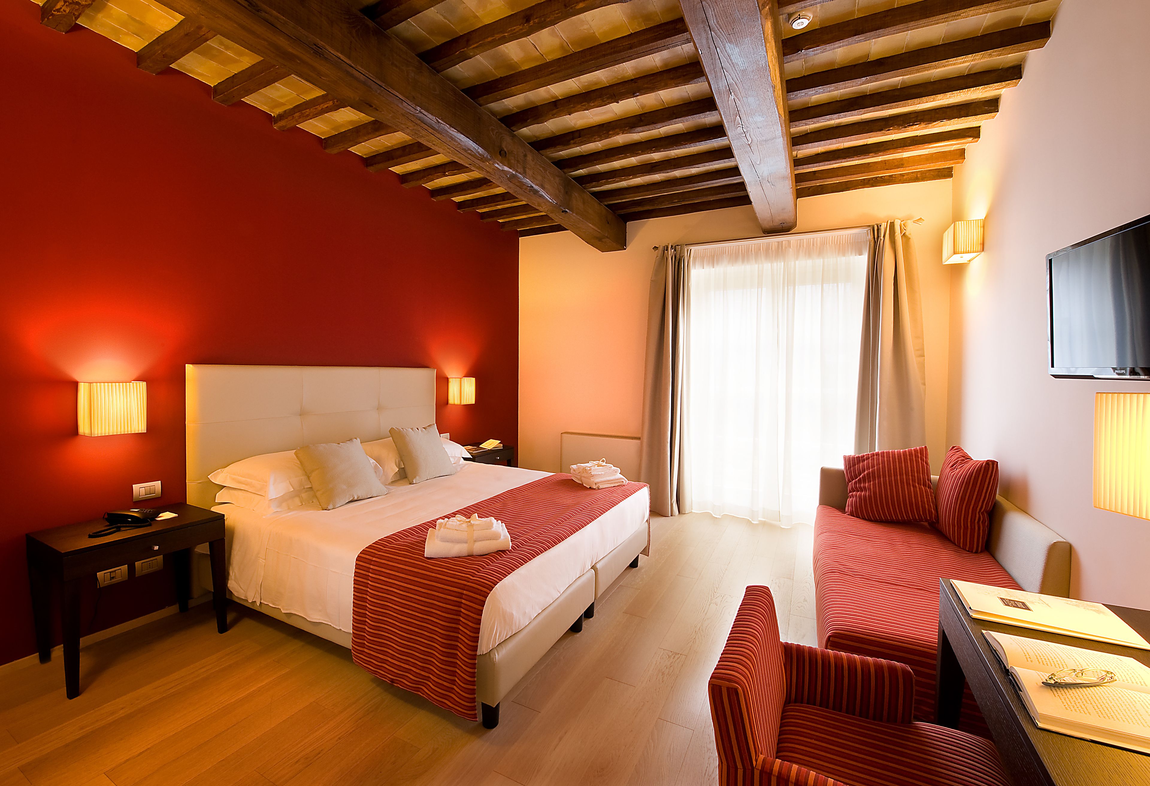 A hotel room with a large bed, two bedside tables, a desk with a chair, a TV, and a window with curtains.