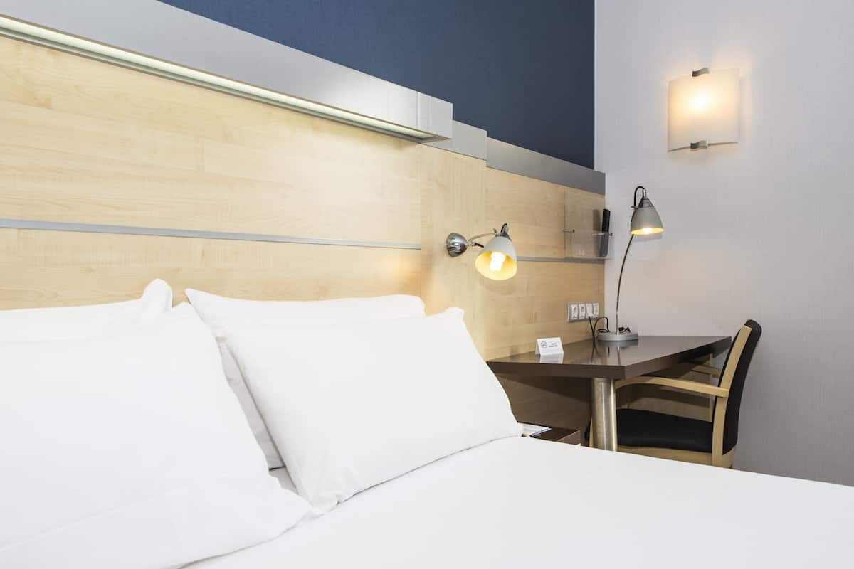 A hotel room with a bed, a desk, and two wall-mounted lamps.