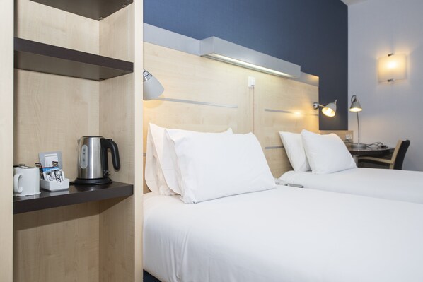 Ein Hotelzimmer mit zwei Betten, einer Kaffeemaschine, einem Wasserkocher und einem Schreibtisch mit Lampe.