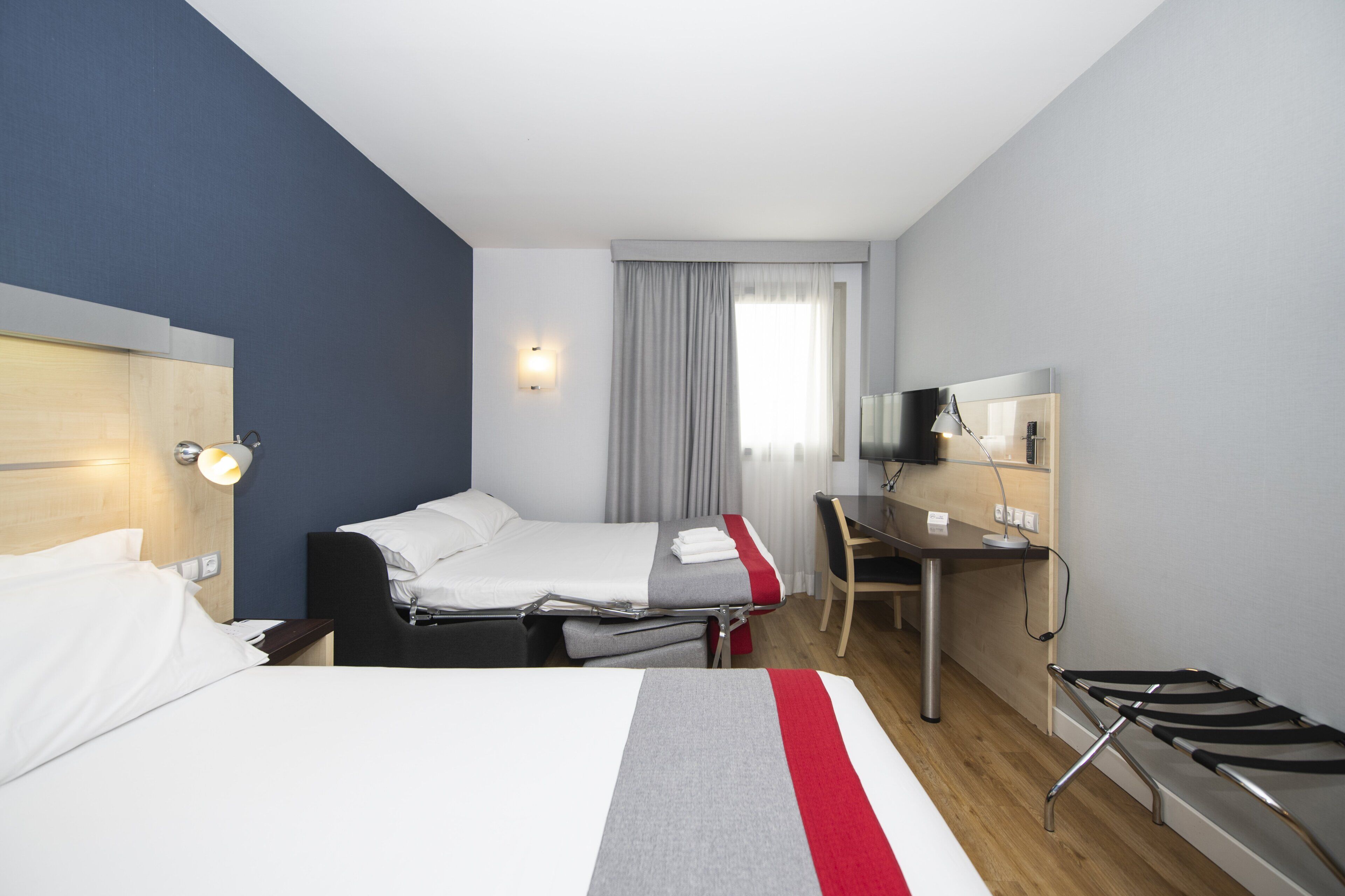 A hotel room with two beds, a desk, a chair, a TV, and a window with curtains.