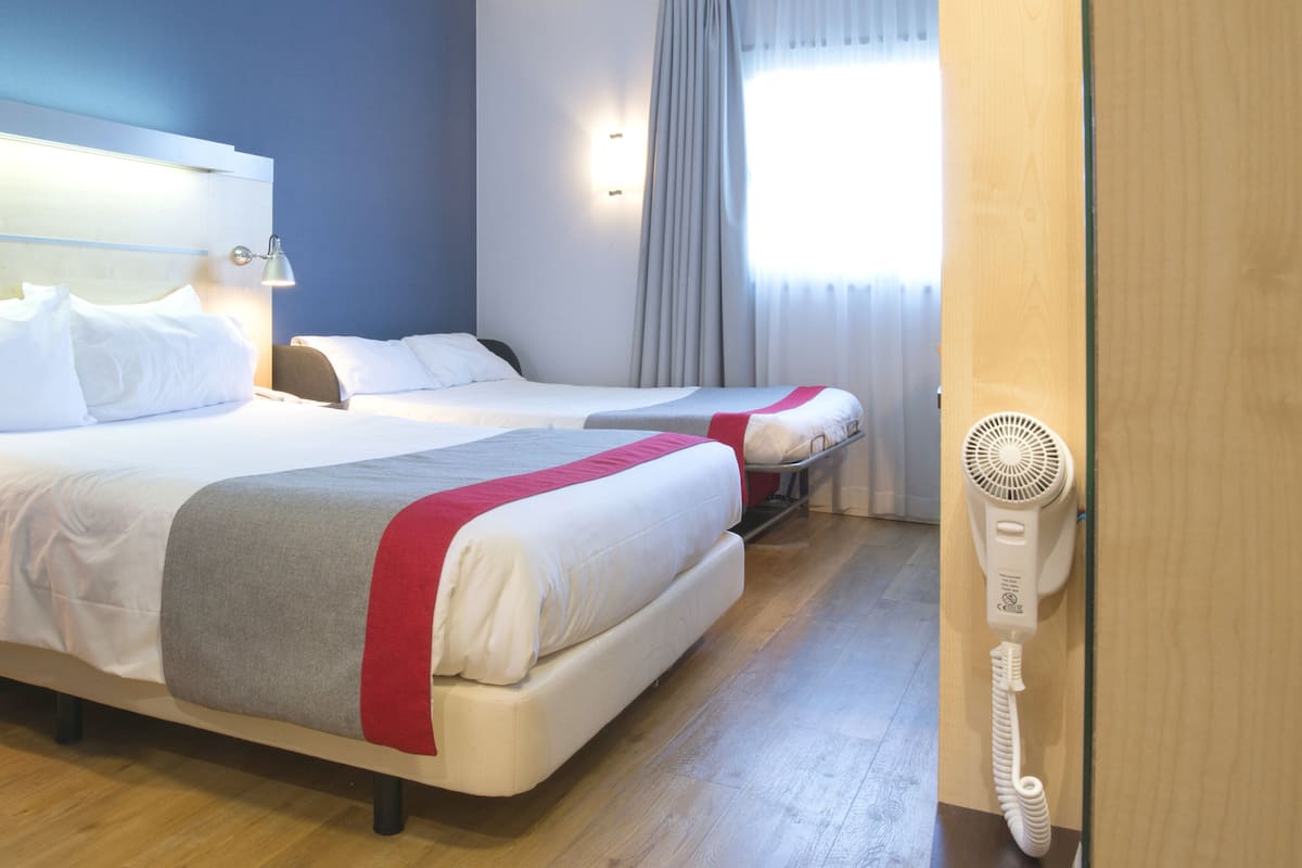 A hotel room with two beds, a bedside table, a lamp, a window with curtains, and a wall-mounted fan.