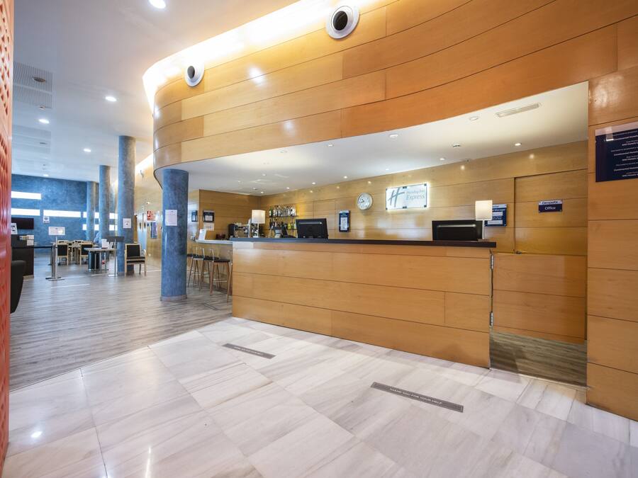 A modern hotel reception area with a wooden reception desk, a digital clock, and a seating area with tables and chairs.