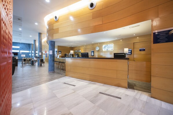 Ein modernes Hotel-Foyer mit einer Holztresenrezeption, einer digitalen Uhr und einer Sitzgelegenheit mit Tischen und Stühlen.